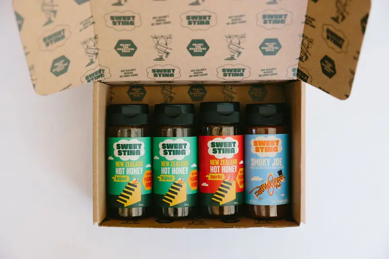 The Sweet Sting gift box open from above with four bottles lying side by side inside — two green Hot Honey, a red Superhot and a blue Smoky Joe — the lid and box interior printed all over with green Sweet Sting cloud logos.