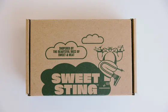 The closed gift box seen from above: kraft cardboard printed in dark green with the Sweet Sting cloud logo, a bee carrying a tray of chillies and a jar, and the line “inspired by the beautiful buzz of sweet and heat”.