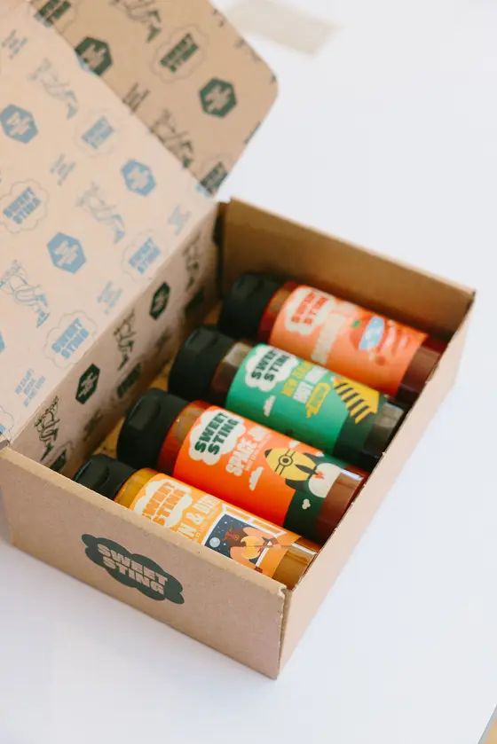 The gift box open at an angle on a white surface, four bottles stacked inside including Space Jam and Down & Dirty Mustard, with the inside of the lid printed in green and blue Sweet Sting logos.